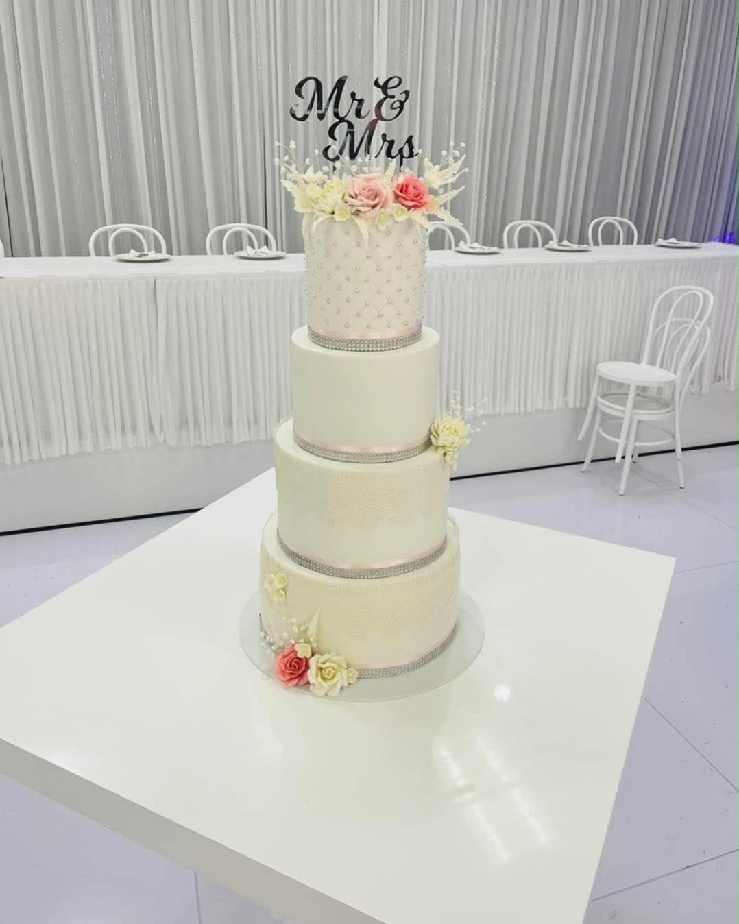 Four tier ivory fondant wedding cake with diamante ribbon borders, pearl dot and quilted fondant details, pink and coral sugar roses and a black Mr & Mrs acrylic topper displayed in a wedding venue